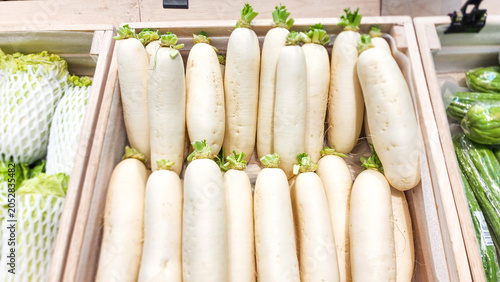 Fresh white daikon radishes with green tops arranged neatly in a wooden crate at a supermarket display counter.