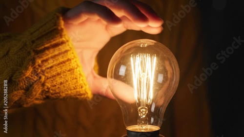 Hand in yellow sweater cupping a glowing vintage Edison light bulb in the dark