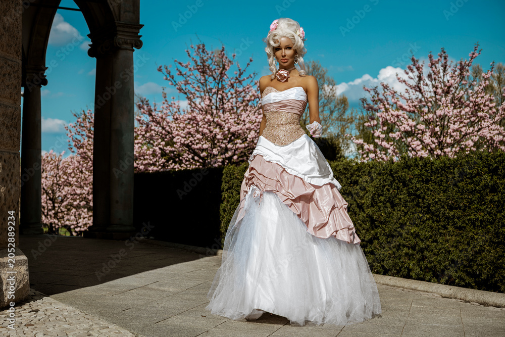 Naklejka premium Gorgeous aristocratic woman in an ornate pink and white baroque ballgown and powdered wig posing in a vibrant spring garden with blooming cherry trees and roses