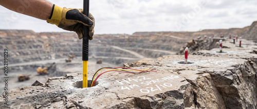 Mining worker inserts a drill rod and connects explosive wires in marked blast holes along the rim of an open-pit quarry, preparing for a controlled rock blasting operation.