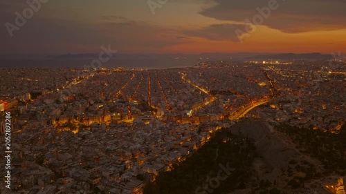 Sweeping aerial view of athens, greece, during twilight, showcasing the sprawling city's illuminated street grid, the dark silhouette of lycabettus hill, and the aegean sea on the horizon