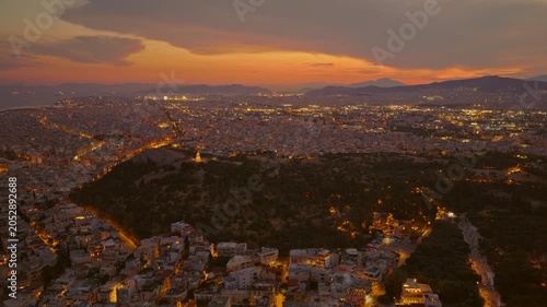 Vast aerial panorama of the city of athens, greece, showcasing the illuminated cityscape and streets under a beautiful orange sunset sky as night approaches with philopappos hill in the foreground