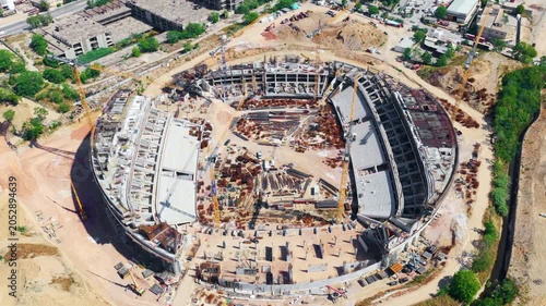 Large-scale construction site of a modern stadium or arena, showing the concrete structure, reinforcement steel, and cranes working on the project, captured from a high aerial perspective