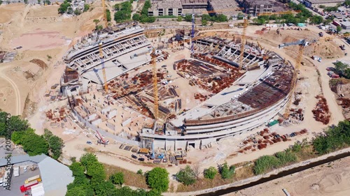Majestic aerial view showing the construction of a large circular stadium, highlighting the intricate network of cranes, concrete structures, and reinforcement bars shaping the future arena