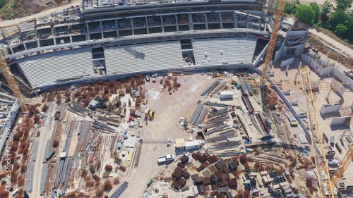 Impressive aerial view showing the massive construction of a new sports stadium, with grandstands taking shape amidst cranes, concrete structures, and building materials scattered across the site