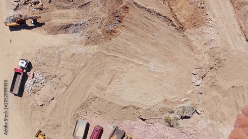 Top-down aerial view of a large yellow excavator digging earth and loading soil into a dump truck at a busy industrial construction site, with other heavy machinery parked nearby