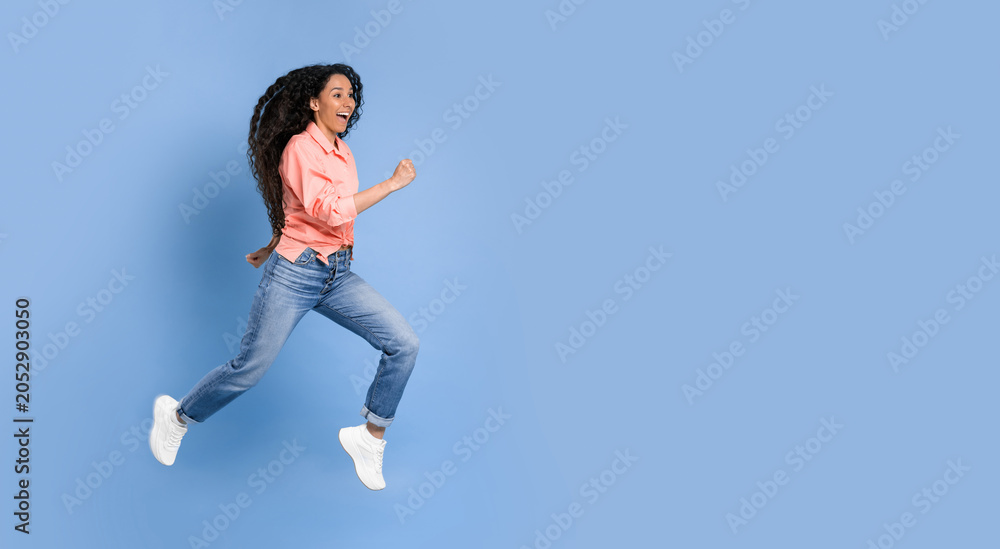 Fototapeta premium A woman with curly hair runs energetically against a solid blue backdrop. She wears a pink shirt and blue jeans, showcasing her movement. Her expression shows joy and enthusiasm while she runs