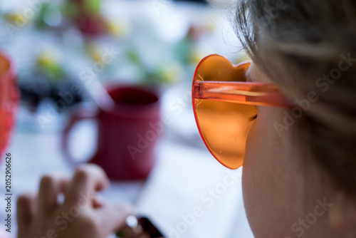 Foto Over-the-shoulder shot of a stylish young girl wearing trendy orange heart sunglasses looking at a brightly lit breakfast table with a red goblet