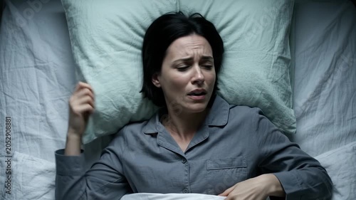 A woman in grey pajamas tosses on a green pillow, clenching her fists in distress. Her painful grimaces convey a clear visual message of vivid nightmares and restless sleep.