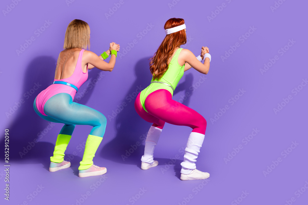 Fototapeta premium Two women in vibrant workout gear performing exercises against a purple background, showcasing a retro fitness style.