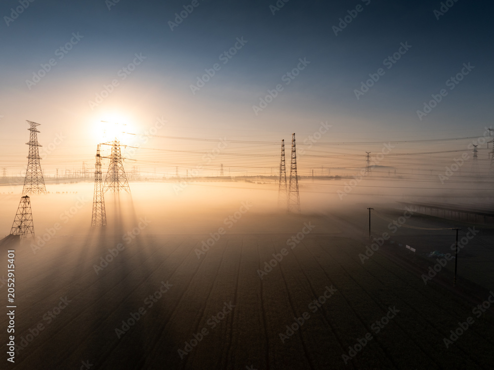 Fototapeta premium Power transmission towers in morning fog with sunlight creating a serene industrial landscape