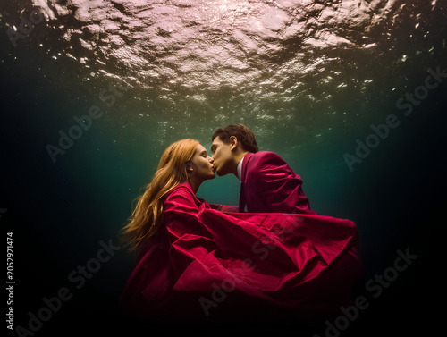The couple kisses while submerged, their hair and clothing flowing upward, surrounded by faint bubbles and light rays piercing the water