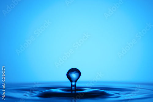 Fotografie Crystal Clear Water Droplet Splashing on Calm Surface Against Vibrant Blue Backg