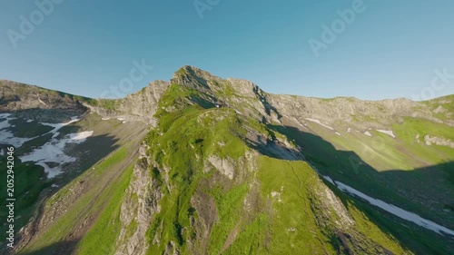 Fast FPV drone footage rushing over a green alpine slope toward rugged rocky peaks, with snow remnants, steep terrain and clear blue sky creating a dynamic summer mountain travel scene