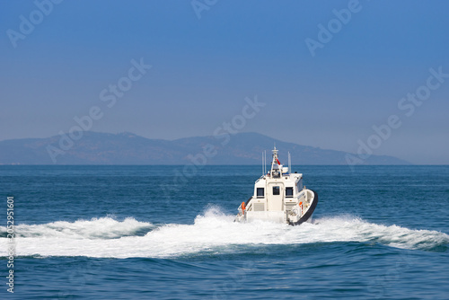 Foto Rear view of a white coast guard patrol boat cruising at high speed on the blue sea water, leaving a white foam wake with mountains in the background