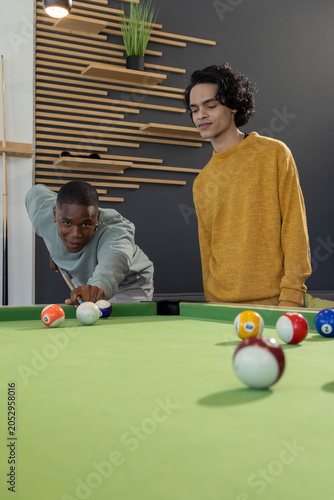 Diverse male friends leaning, taking shot on green pool table in modern lounge with cue