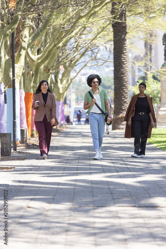 Three friends walking down tiled promenade under canopy, carrying coffee cup and crossbody bag