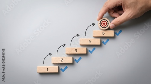 A hand places a bullseye on the fifth step of a numbered staircase, symbolizing the achievement of a goal. The steps are marked 1 through 5, with checkmarks on each step.