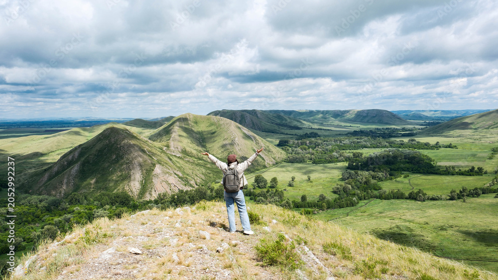 Fototapeta premium Alone backpacker woman stands on a rocky hillside overlooking a wide green valley with rolling mountains, scattered trees, and a cloudy sky in the background