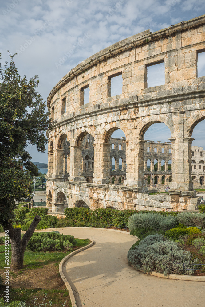 Obraz premium The Pula Arena, a Roman amphitheatre. The historic stone arches and walls show the grandeur of ancient architecture. A winding path lead towards the colossal structure. Pula, Croatia.
