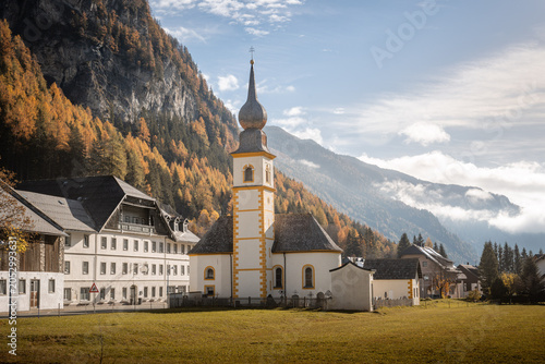 Fotografía White and yellow decorated church with an onion dome steeple standing prominently in a green field, surrounded by traditional buildings and autumn trees, Tweng, Austria