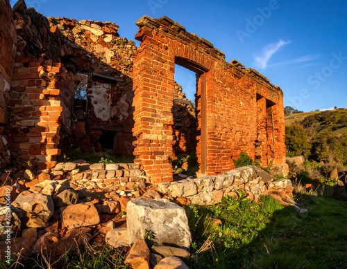 Crumbling brick structure bathed in golden hour light, revealing open doorways and stone foundation. Greenery surrounds
