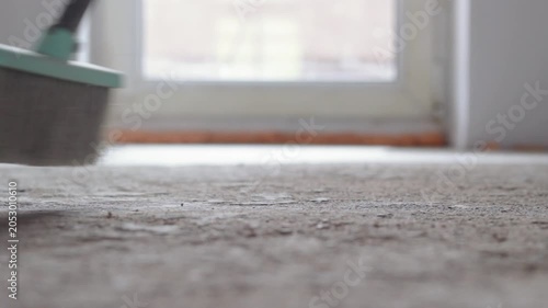 Close-up of a dusty concrete floor being swept clean with a sweeping broom in an empty indoor space. Daylight softly highlights the dust particles. Blurry background with a window