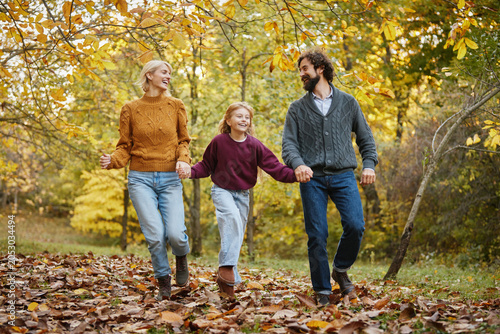A cheerful family walks hand in hand through a park filled with fallen leaves. They smile and laugh together under the vibrant autumn trees. A sense of happiness fills the scene during the weekend.