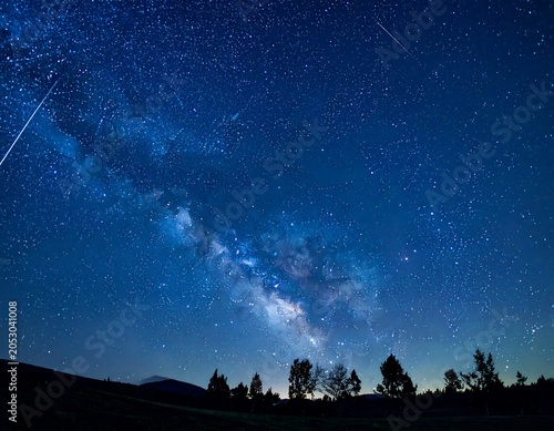 Deep blue night sky, a breathtaking cosmic display featuring the Milky Way, meteor streaks, and a silhouetted treeline