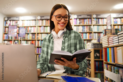 Foto Positive female student reading book in front of laptop in university library si