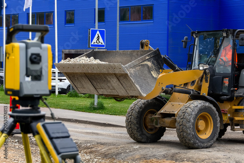Wheel loader with crushed stone bucket and blurred total station at road construction site