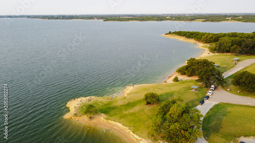 Lakeside park peninsula Lake Lavon shoreline near Princeton with grassy curves, sandy coves, trees, vehicles along road beside shelters. Mixed terrain patterns, water gradients aerial view, Texas