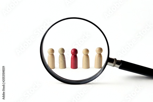 A magnifying glass focusing on a red wooden peg doll standing out in a row of natural figures, isolated on white background, concept of talent scouting, recruitment, and business auditing.