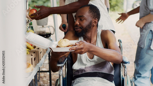Foto Free meals and fruits are given to a wheelchair-bound African American man in need, with handicapped, poor and homeless people receive provisions from a non-profit food drive Close-up, tripod shot