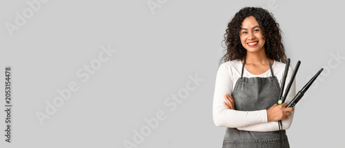 Beautiful young happy African-American hairdresser with hair straightener and...
