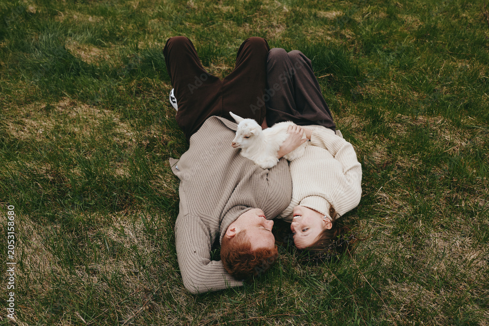 Fototapeta premium A couple lies on a grassy field, cuddling a white kid goat between them, sharing a calm moment in the open air. A gentle, lifestyle scene focused on family bonding and rural simplicity.