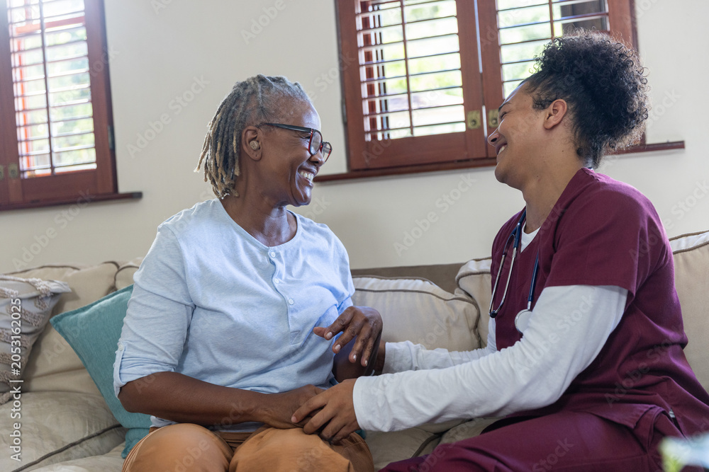 Fototapeta premium Senior African American woman sitting on sofa holding hands with female nurse wearing stethoscope