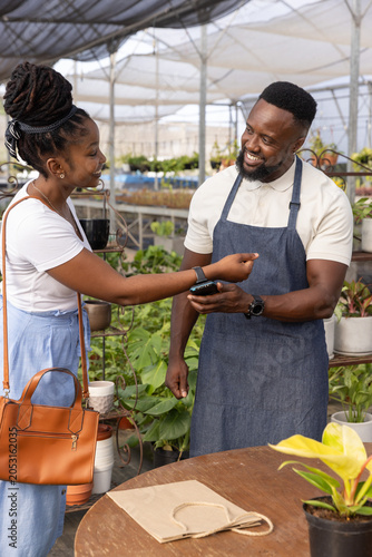African American customer showing phone to nursery staff in denim apron while carrying tan handbag