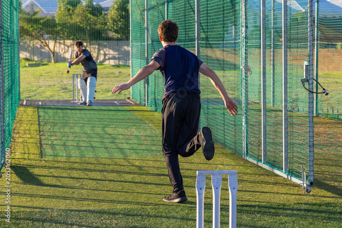 Two men practicing cricket in net on turf wearing sportswear  holding bat  ball and white stumps