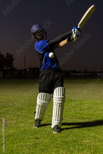 Cricket batter is swinging wooden bat with white ball on grass field at dusk under lights