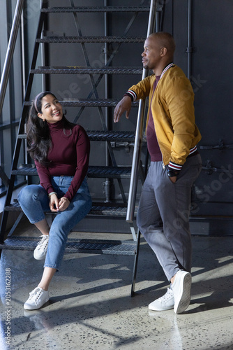 Diverse couple chatting on metal staircase in industrial loft by window, wearing maroon tops