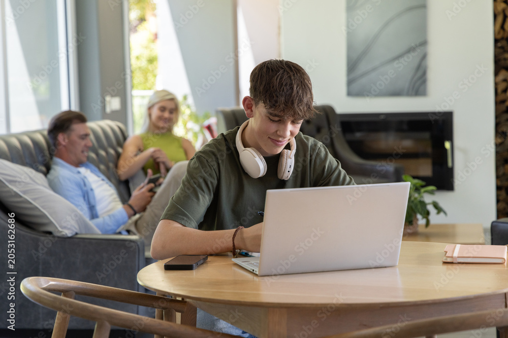 Obraz premium Diverse family using smartphones on sofa as youth male working on laptop at living room table
