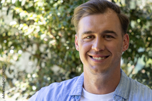 Man smiling and facing camera in garden, wearing light blue button-up shirt over white T-shirt