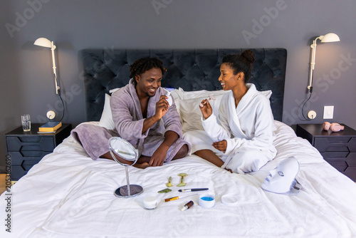 African American couple wearing robes sitting on bed, applying skincare with rollers and LED mask