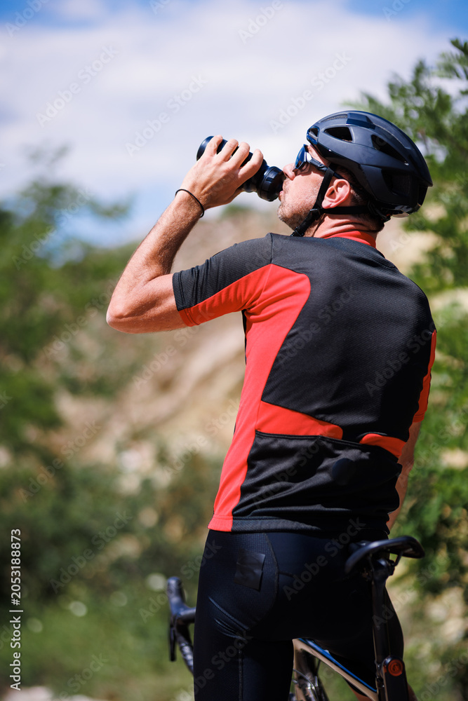 Fototapeta premium Cyclist drinks water while resting near mountain trail during sunny day in the afternoon with bike nearby