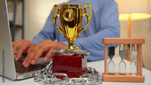Bild auf Leinwand A freelancer works on a laptop beside a trophy chained tight and an hourglass, symbolizing how achievements can feel restricted by time, pressure, deadlines, sacrifice, and the burden of expectations