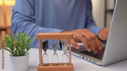 Tableau sur toile A person works at a laptop with an hourglass beside them, symbolizing time pressure, deadline-driven productivity, effective time management, prioritization, and maintaining focus on results