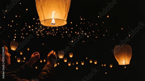 Hands hold lit lantern with warm flame. Close-up shows careful lighting action. Person releases lantern into night sky. Calm expression visible on face. Many lanterns float with soft glows