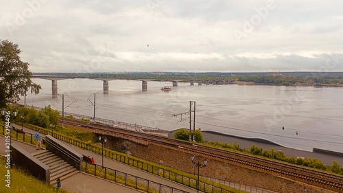 Urban Riverfront Landscape with Concrete Bridge, Train Tracks, and Pedestrian Walkway under Cloudy Sky