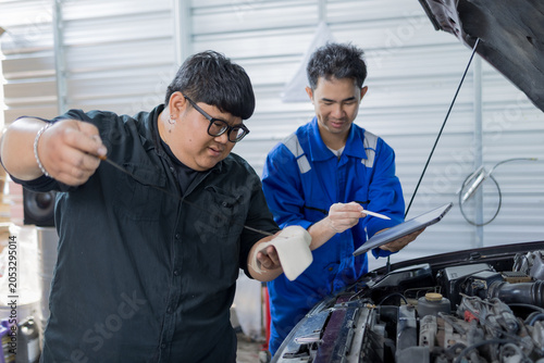 Two mechanics examine a car engine together. One holds a clipboard, the other a laptop. They focus on the engine's components closely. Their expressions show concentration and teamwork
